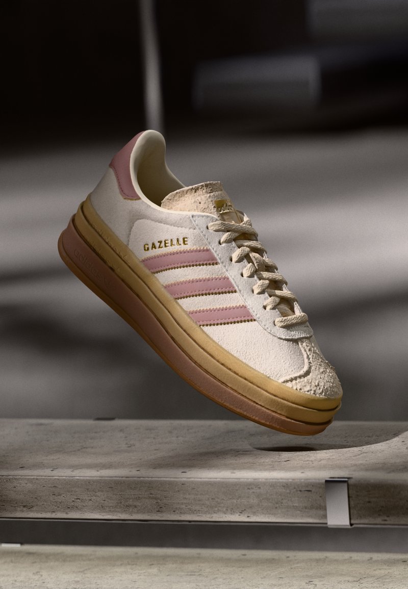 Shoe de camurça branca com detalhes em rosa, apresentando três riscas e um logo dourado. Biqueira texturizada e sola de borracha castanha. Design clássico.