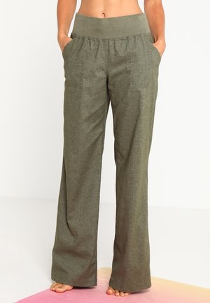 Osoba stojící bosá na růžové a žluté podložce na jógu, oblečená do kalhot s vysokým pasem, širokými nohavicemi a kapsami olivově zelené barvy.