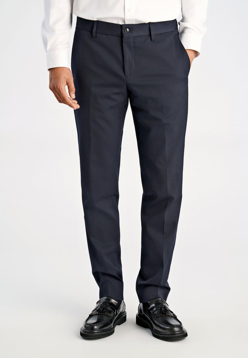 Marineblaue Anzughose in einem schlanken, maßgeschneiderten Design mit flacher Vorderseite und Bügeldetails; kombiniert mit schwarzen Lederschuhen und einem weißen Hemd.