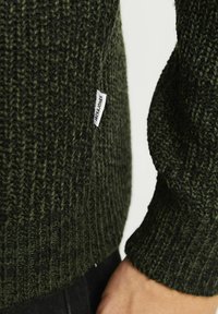 Dunkelgrüner gestrickter Pullover mit gerippter Textur, versehen mit einem Etikett an der Seite. Das Design zeigt ein weiches, dickes Strickmuster.