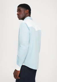 Camicia a maniche lunghe azzurro chiaro con yoke posteriore bianco, polsini con bottoni e orlo leggermente curvato, realizzata in un tessuto morbido.