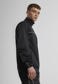 Black zip-up jacket with a high collar, white piping details, and elastic cuffs. Made of smooth fabric. Model shown from the side.