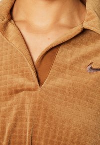 Braun strukturierter Quarter-Zip-Oberteil mit einem karierten Muster, das einen kontrastierenden braunen Blenden und ein dezentes gesticktes Logo auf der Brust aufweist.