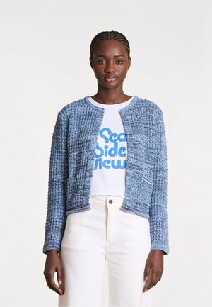 Femme aux cheveux tressés portant un cardigan bleu texturé ouvert sur un t-shirt blanc à motif et un pantalon blanc taille haute.