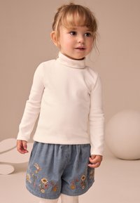 White long-sleeve turtleneck, paired with blue corduroy shorts featuring floral embroidery. Light background, soft textures.