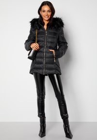 Svart vadderad jacka med huva i faux päls, dragkedjestängning och sidfickor. Kombineras med eleganta svarta leggings och spetsiga ankelboots.