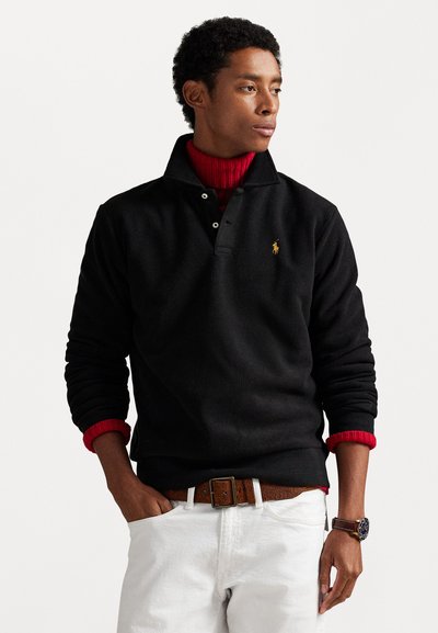 Sort fleece-pullover med krave, der har et logo på venstre bryst. Båret over en rød sweater, kombineret med hvide bukser og et brunt bælte.