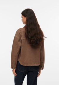 Καφέ τζιν μπουφάν με χαλαρή εφαρμογή, με κυρτή μπάσκα, μανίκια με κουμπιά και ραμμένες ραφές. Φορεμένο πάνω από σκούρο μπλε τζιν παντελόνια.