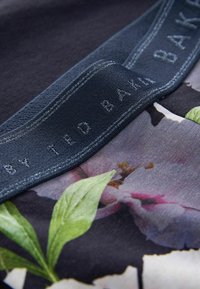 Blumenmusterstoff mit dunklem Hintergrund, der auffällige grüne Blätter zeigt, sowie ein strukturierter blauer Aufnäher mit dem eingeprägten Schriftzug "TED BAKER" in Silber.