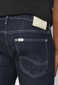 Ciemnoniebieskie dżinsy z denimu z kontrastującymi białymi przeszyciami, etykietą na tylnej kieszonce oraz przypinką przypominającą skórę z wytłoczonym napisem "Lee".