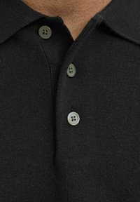 Közeli kép egy fekete, textúrált pólóról, három szürke gombbal a gombolás mentén és egy hajtott gallérral.