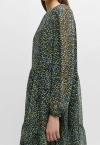 Blumenkleid aus transparentem Stoff, mit einer Mischung aus schwarzen, blauen und grünen Blumen. Lange Ärmel, lockere Passform und geraffte Taillendetails.