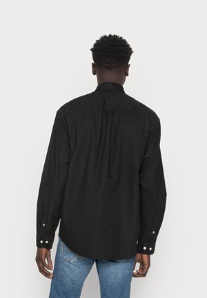 Chemise noire à manches longues en coton, avec un col boutonné et un pli au dos. Associée à un jean en denim bleu clair.