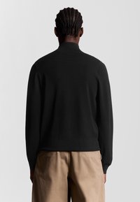 Sort cardigan med høj krave og lange ærmer, lavet af et glat stof, parret med beige bukser. Set fra ryggen.