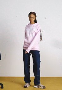 Rosa hettegenser med hvit adidas-logo, mørkeblå jeans og mønstrede sneakers. Modellen har lange fletter og står mot en nøytral bakgrunn.