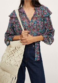 Blusa floral con mangas acampanadas, de tela multicolor, combinada con un bolso de macramé crema, que presenta patrones intrincados y flecos.