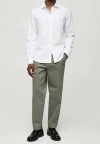 Camisa blanca de manga larga con botones, combinada con pantalones verdes oliva claros y zapatos de vestir negros. La camisa presenta un cuello clásico y un diseño ajustado.