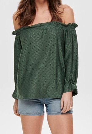 Kvinde iført en grøn off-shoulder langærmet bluse med huldetaljer og lyseblå denimshorts mod en ensfarvet baggrund.