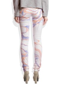 Femme portant un pantalon slim à motif marbré pastel avec des nuances d'orange et de violet, assorti à des chaussures à talons haut beiges et un pull gris.