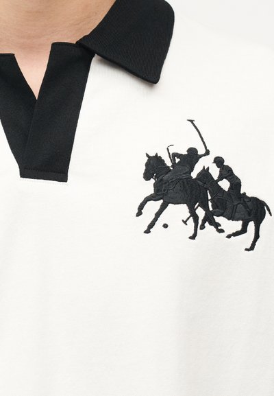 Δύο παίκτες πόλο πάνω σε άλογα με υψωμένα ρόπαλα κυνηγούν μια μπάλα κεντημένη με μαύρο χρώμα σε λευκό πουκάμισο με μαύρο γιακά.