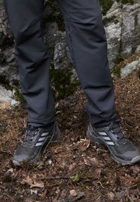 Czarne buty trekkingowe z szarymi akcentami i trzema białymi paskami, w połączeniu z czarnymi spodniami. Buty mają teksturowaną powierzchnię i wzmocniony nos.