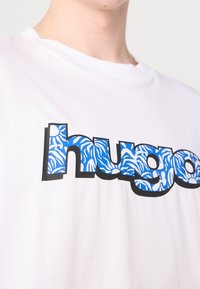 Bílý bavlněný tričko s výrazným modrým nápisem "hugo" a květinovým vzorem. Černý obrys kolem písmen přidává kontrast.