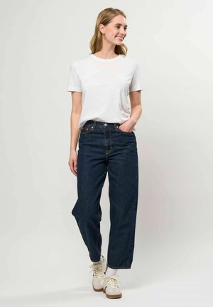 Jonge vrouw staat, glimlacht, draagt een wit T-shirt, donkerblauwe high-waisted jeans, witte sokken en beige sneakers, tegen een effen achtergrond.