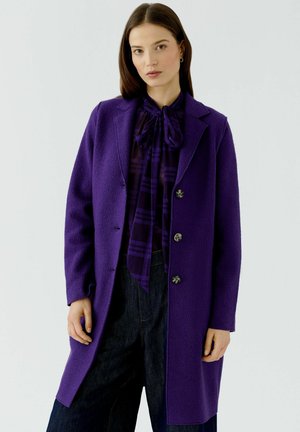 Femme aux cheveux raides et foncés portant un manteau violet boutonné par-dessus un chemisier assorti et un pantalon foncé, debout devant un fond clair uni.