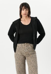 Zwarte zip-up hoodie met een capuchon, gecombineerd met een zwarte tanktop en een leopardprint high-waisted jeans met een relaxte pasvorm en zakken.