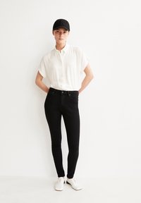 Camisa de botones de manga corta blanca, jeans ajustados negros y zapatos blancos con cordones. El modelo lleva una gorra negra con un emblema. Fondo liso.