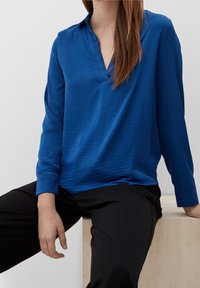 Blusa blu a maniche lunghe con colletto e tessuto testurizzato, abbinata a pantaloni neri. Presenta una vestibilità rilassata e maniche leggermente larghe.
