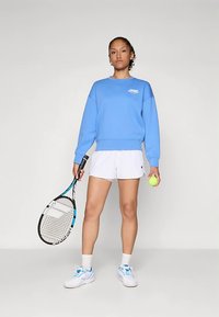 Γυναίκα που στέκεται κρατώντας μια ρακέτα τένις και μια μπάλα, φορώντας μια μπλε φούτερ, λευκά σορτς, λευκές κάλτσες και λευκά παπούτσια τένις σε απλό φόντο.