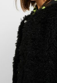 Manteau noir texturé avec une finition duveteuse, doté d'une doublure verte à motifs et de boutons argentés. Le design est épais et ample, mettant l'accent sur la chaleur.