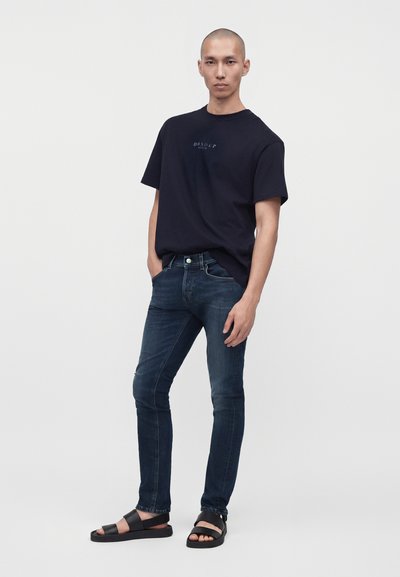 Άνδρας φοράει μαύρο T-shirt, σκουρό μπλε τζιν και μαύρα σανδάλια, στέκεται με το ένα χέρι στην τσέπη μπροστά σε απλό λευκό φόντο.