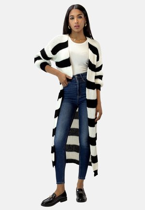 Black and white striped long cardigan with ribbed texture, worn over a fitted white top and high-waisted blue jeans. Black loafers.