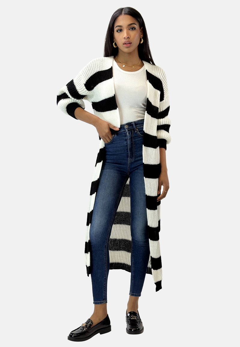 Langer schwarz-weißer gestreifter Cardigan mit gerippter Textur, getragen über einem eng anliegenden weißen Oberteil und hochgeschnittenen blauen Jeans. Schwarze Slipper.