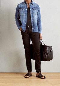 Dżinsowa koszula z metalowymi guzikami, czarne spodnie o kroju slim oraz czarne klapki. Niesiona jest czarna skórzana torba podróżna. Widać drewnianą podłogę.