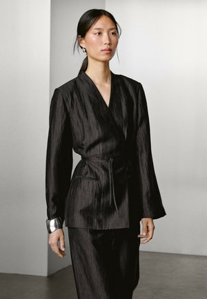 Une femme portant une veste enveloppante noire texturée et un pantalon assorti marche avec assurance, mettant en avant une mode simple mais élégante dans un cadre moderne.