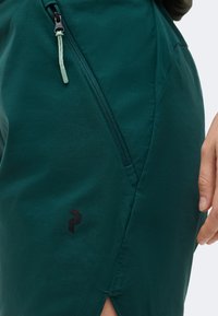 Τυρκουάζ φούστα με διαγώνια τσέπη με φερμουάρ, μικρή σχισμή στο τελείωμα και μαύρο γεωμετρικό λογότυπο κοντά στην τσέπη· χέρι που ακουμπάει στο πλάι.