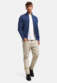 Jonge man draagt een blauwe trui met rits, wit T-shirt, beige broek en zwarte schoenen, staand met één hand in zijn zak.