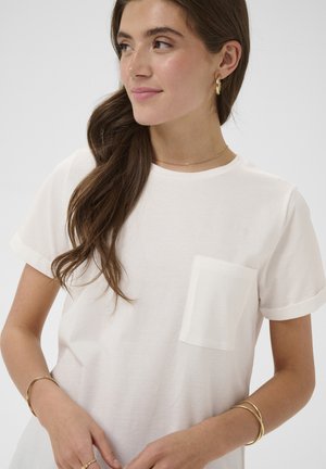 Mlada ženska z dolgimi rjavimi lasmi v beli majici s kratkimi rokavi in žepom, ki nosi zlate obročaste uhane, ogrlico in več zlatih zapestnic, gleda desno.