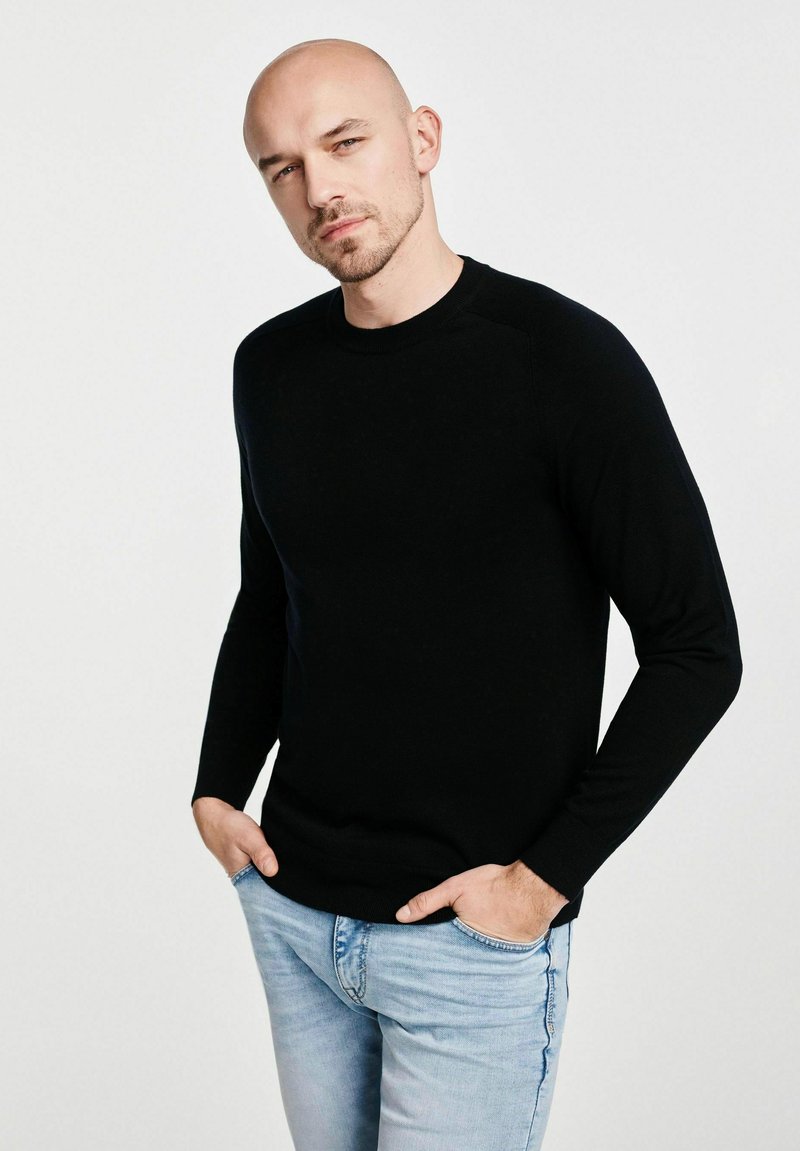 Czarny sweter z długim rękawem, wykonany z gładkiego materiału, z okrągłym dekoltem i ściągaczami przy mankietach, noszony z jasnoniebieskimi dżinsami.