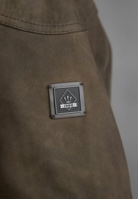 Smeđi materijal glatke teksture, s crno-srebrnim logotipom u obliku trokuta s godinom "1982" na dnu.
