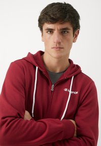 Červená mikina na zips vyrobená z bavlneného zmesi, s bielymi šnúrkami, logom na hrudi a rebrovanými manžetami s hladkou textúrou.