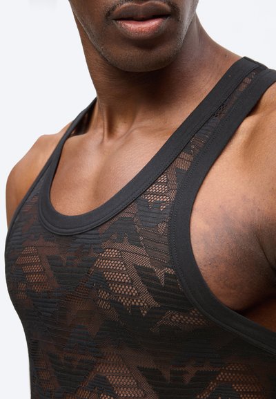 Κοντινό πλάνο ενός άνδρα που φοράει μαύρο δίχτυ μπλουζάκι με γεωμετρικά σχέδια, εστιάζοντας στη γραμμή του λαιμού και το πάνω μέρος του στήθους.