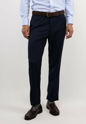 Hombre con pantalones de vestir azul marino, camisa azul claro a rayas metida dentro, cinturón marrón, calcetines grises y mocasines de cuero marrón oscuro.