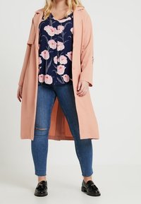 Cappotto lungo rosa chiaro con colletto e bottoni, indossato sopra una blusa floreale blu navy con fiori rosa, abbinato a jeans blu e scarpe nere.