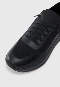 Svart sportsko med mesh-overdel, gummidetaljer og snøringsdetaljer. Merkevaren er subtilt vist på siden.