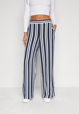 Vrouw draagt marineblauwe, wijde broek met hoge taille en verticale witte strepen, gecombineerd met witte sneakers en een wit crop top met lange mouwen.