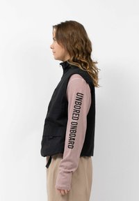Schwarze ärmellose Weste mit Reißverschluss und Fronttaschen, getragen über einem langärmeligen hellrosa Oberteil mit schwarzem Text entlang des Ärmels.